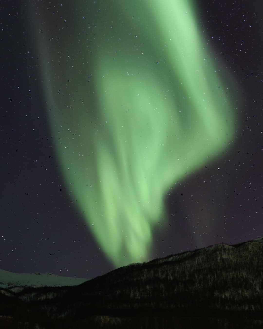A bright green aurora curtain above a dark mountain under a starry sky near Tromsø, Northern Norway.